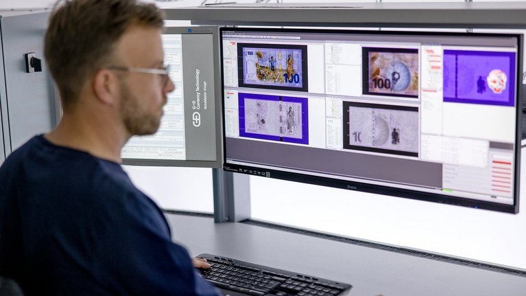 A person is working at a desk with two large computer monitors displaying images of currency notes
